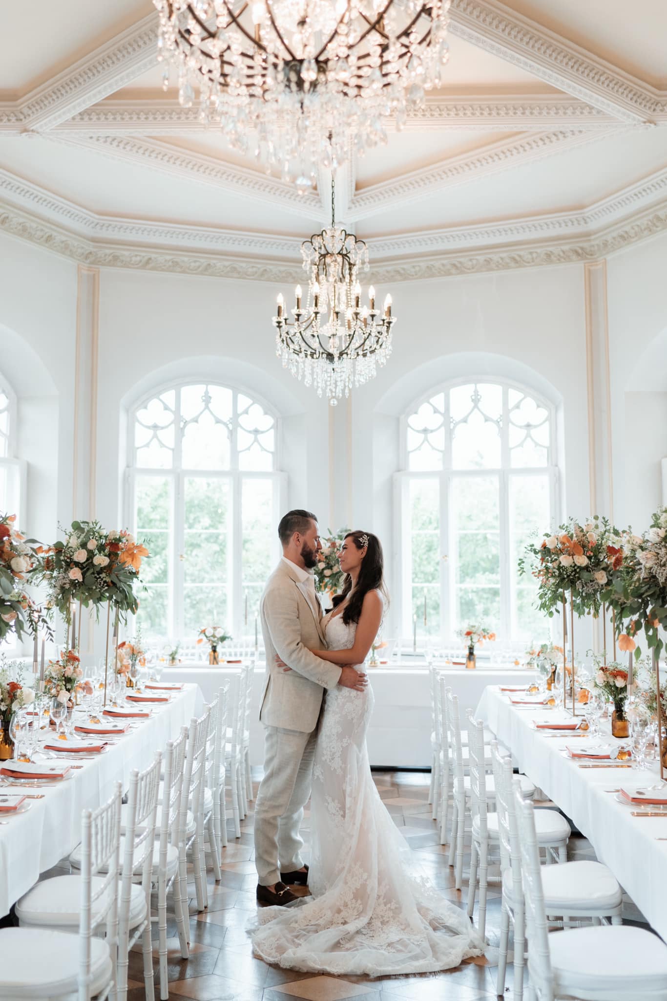 Hochzeitspaar in weiß steht in weiß eingerichtetem Raum im Schloss Gamehl mit Konleuchter.