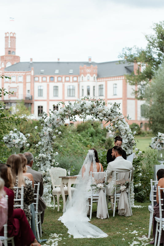 Hochzeitspaar sitzt vor Traubogen und lauscht der Rednerin, im Hintergrund ist das Schloss Gamehl zu sehen.