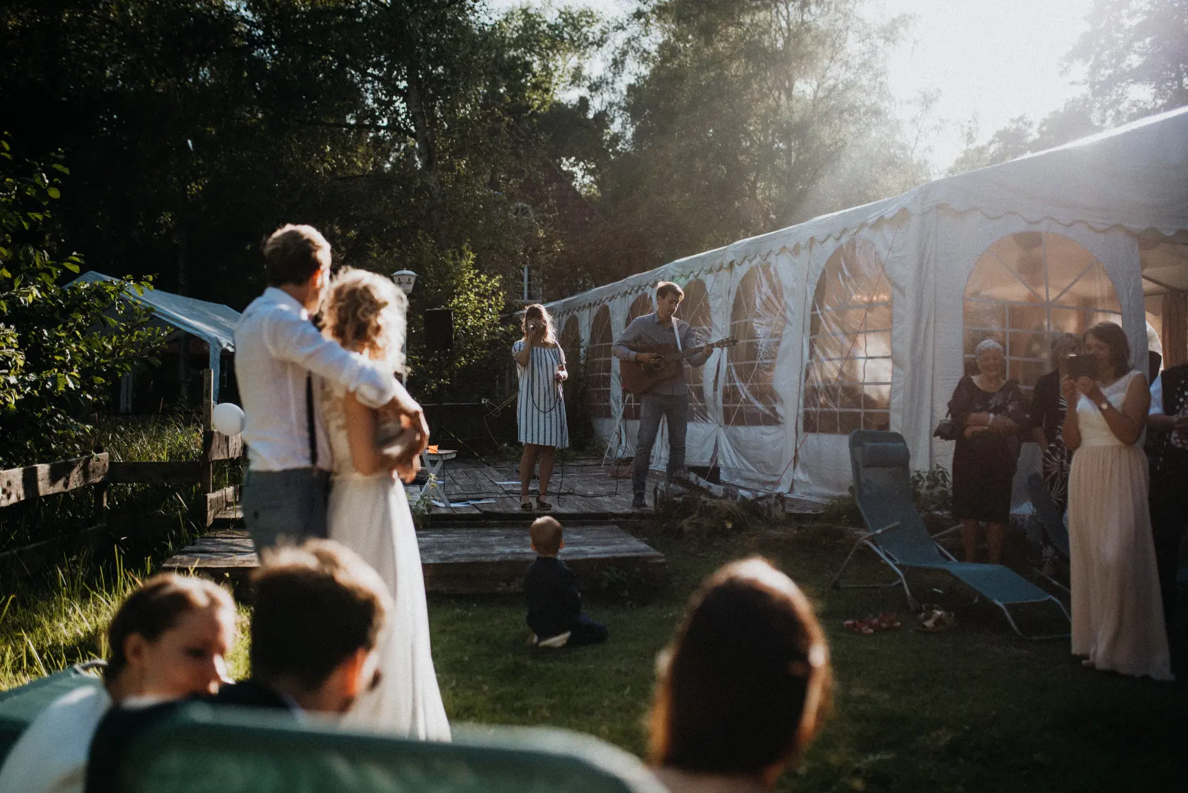 Das Hochzeitspaar lauscht der Musik von ihrer Hochzeitssängerin und einem Gitarristen im Restaurant Müggenbusch in Lübeck bei strahlendem Sonnenschein.